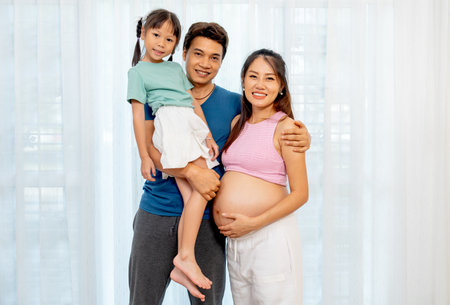 Asian father carry little girl and stand beside pregnant mother in front of white curtain with smiling and look at camera.の写真素材
