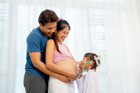 Asian father hug pregnant mother in her back and little girl use stethoscope toy to play with belly of mother in front of white curtain in their house.の写真素材