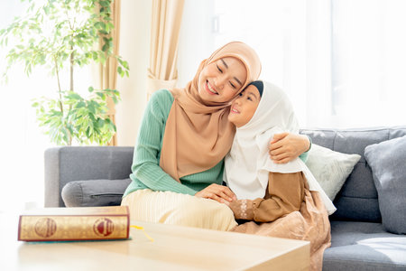 Asian muslim mother sit on sofa beside of her daughter and hug her child they also smiling in concept of happiness muslim family at home.の写真素材