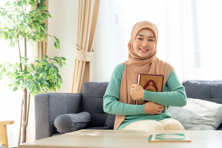 Asian beautiful muslim woman sit on sofa in her house and hug or hold in her chest with the book relate to her religion or islamic teaching book and look at the camera also smile with happiness.の写真素材