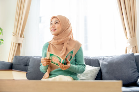 Asian beautiful muslim woman sit on sofa in her house and read the book relate to her religion also look to her upper right side with smiling.の写真素材