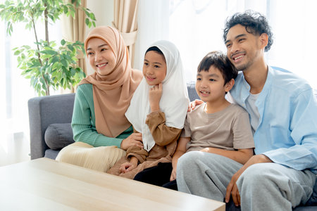 Asian muslim family compose of young mother, father, daughter and son hug and stay together on sofa in living room of their house and look forward and right direction of image with smiling.の写真素材
