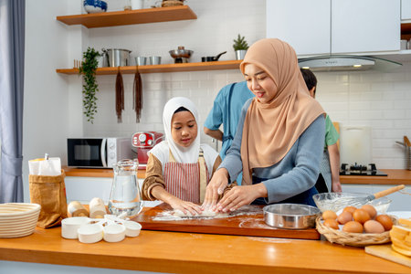 Young Asian muslim mother help and teach her daughter to threshing flour by hands on wooden tray for making the bakery with father and son also cook in the back in the kitchen of their house.の写真素材