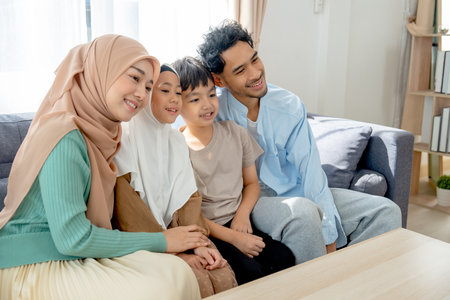 Asian muslim family compose of young mother, father, daughter and son hug and stay together on sofa in living room of their house and look forward and left direction of image with smiling.の写真素材