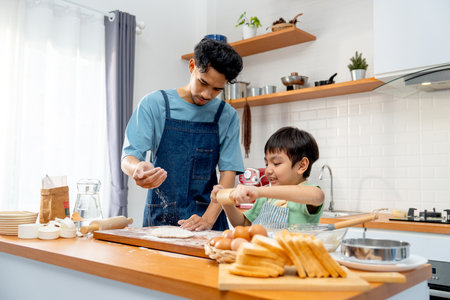 Wide shot of young Asian father pour cooking flour on wooden tray during teaching his son to thresh flour for making bakery in kitchen of their house.の写真素材