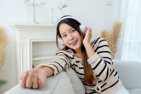 Close up and portrait of young lovely Asian woman with headphone sit and lean on sofa and look at camera with smiling in living room of her house.の写真素材