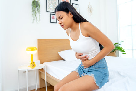 Side view of Asian woman sit on bed and express of stomach ache by use two hands to hold her stomach in bedroom.の写真素材