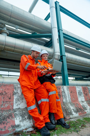 Vertical image of two petrochemical or gas factory workers or engineers man and woman discuss about work using laptop and sit near pipeline in factory workplace.の写真素材