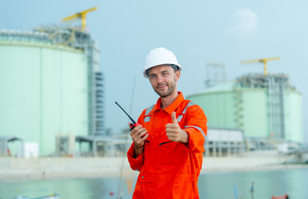 Petrochemical or gas factory worker hold walkie talkie and show thumbs up to camera and stand in...