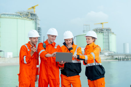 Group of petrochemical or gas factory workers men and woman or technician stand in front of factory tank near the bay and work together using laptop.の写真素材