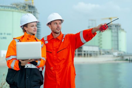 Petrochemical or gas factory workers man and woman work together with one hold laptop and look at direction of man point to left side in front of factory tank near the bay.の写真素材