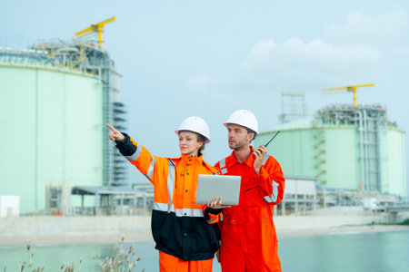 Petrochemical or gas factory workers man and woman work together with one hold laptop and point to ride side and man hold walkie talkie stay in front of factory tank near the bay.の写真素材