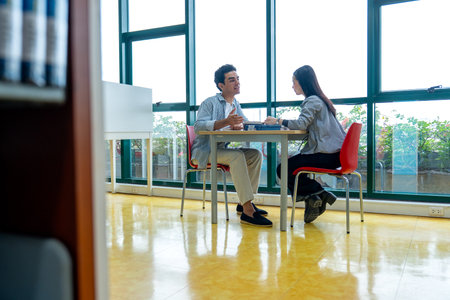 Side view and wide shot Hispanic man sit and discuss with Asian woman at table in public library in front of glass window with day light.の写真素材