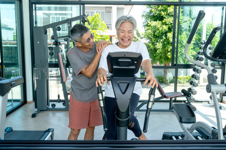 Asian senior man stand beside and hug senior woman during use stationary bike to exercise in sport...