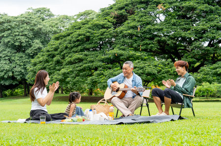 Side view of Asian little girl sit on mat to picnic during grandfather play the music using guitar...