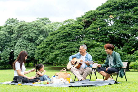 Asian little girl sit on mat to picnic during grandfather play the music using guitar and her father...