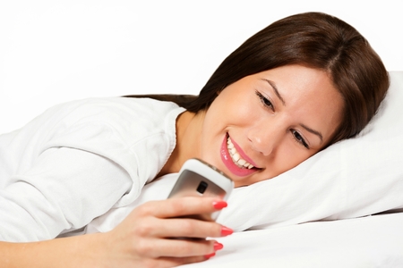 Happy woman lying on bed smiling while reading a text message on mobile phone.の写真素材