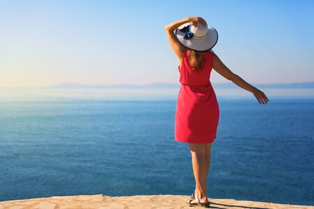 Young woman, wearing white hat and red dress, enjoying sea view.の写真素材