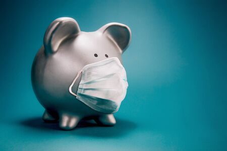 Close up of piggy bank, wearing protective face mask, isolated on blue background. Money saving concept in time of coronavirus pandemic.の写真素材