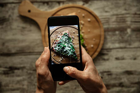 Female hands holding a smartphone and taking picture of a fresh vegan pizza slice.の写真素材