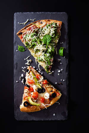 Slices of hot pizza with prosciutto, rocket salad, vegetables, tomatoes, and cheese on blackboard...