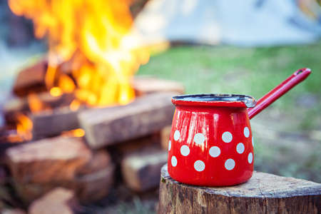 Close up of red coffee pot on top of wood stump, with campfire and camping tent in the background.の写真素材