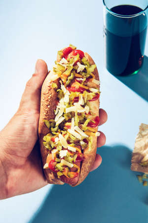 Close up of male hand holding freshly served hot dog with cheese, pickles, ketchup and mustard topping, above fast food restaurant table.の写真素材