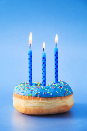 Close up of a donut with blue icing and sprinkles, with lit birthday candles on top of it, isolated against blue background.の写真素材