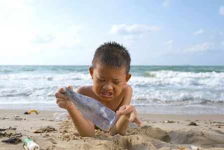Child lay down on the beach and picks up dirty bottles at the beach. A boy picks up dirty plastic bottles at the beach.の写真素材