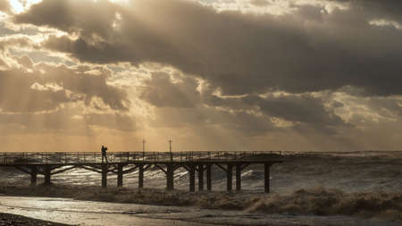 man on a pier taking a photo of sunset cloudy sky and stormy sea