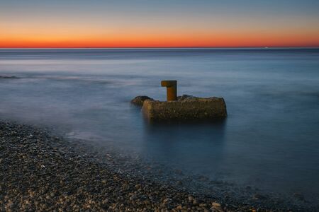 Abandoned sea bollard on rocky beach, with orange sunset sky 