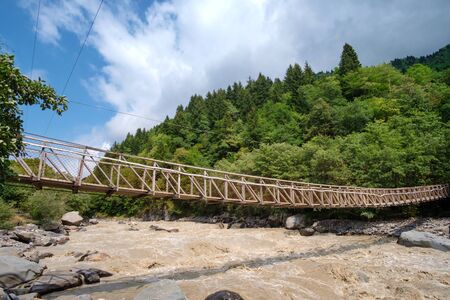 Hanged rope bridge over the fast muddy river with green forest background 