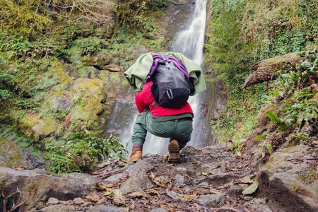 Hiker tourist with backpack is kneeling and taking photo of waterfall at autumn forestの写真素材