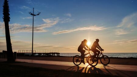 Silhouettes of two young man riding on bikes in sea side park at sunset