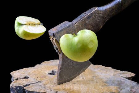 Rusty axe slicing an green apple in half on the wooden board, black backgroundの写真素材