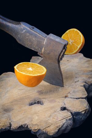 Rusty axe slicing an orange in half on the wooden board, black backgroundの写真素材