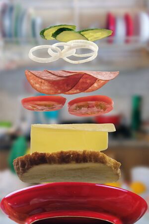 Flying layers of sandwich with ham, cheese, vegetables over the red plate with blurred kitchen background. Breakfast food concept, selective focusの写真素材