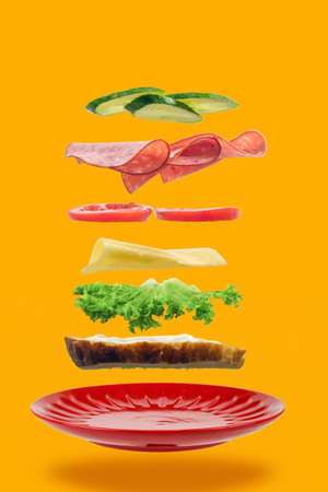 Flying layers of sandwich with ham, cheese and vegetables over the red plate on yellow background. Breakfast food conceptの写真素材