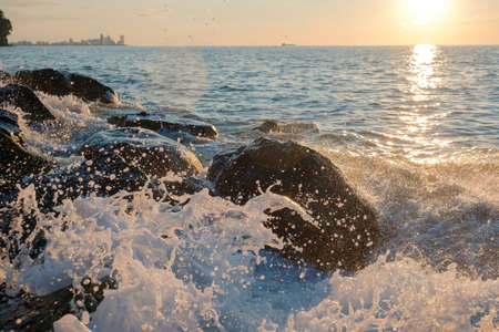 Sea waves crushing over the big rocks with beautiful sunset sky and city line on the background