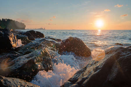 Sea waves crushing over the big rocks with beautiful sunset sky and city line on the backgroundの写真素材