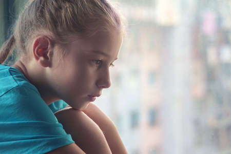 A brown eyed sad girl sitting on the window sill  with blurred outdoor background