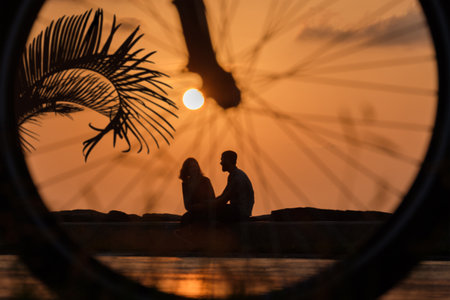 Silhouettes of the couple relaxing at seaside park at sunset, view through the blurred bike wheel,...