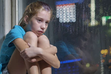A brown eyed girl in blue t-shirt sitting on the window sill behind a foggy window with blurred night backgroundの写真素材