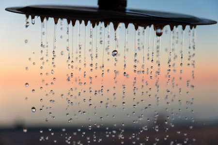 Water drops pouring from the outdoor beach shower head on blurred sunset seascape background, selective focusの写真素材
