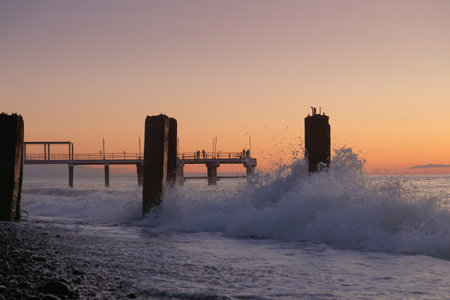 A splashing waves and pier on a rocky beach, sunset backgroundの写真素材