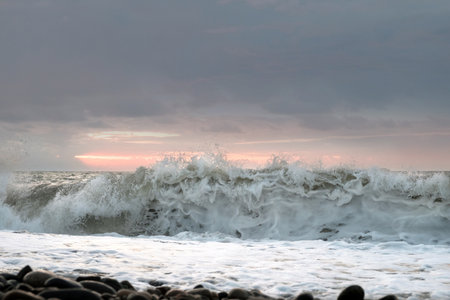 Big foamy splashing sea wave crushing to the pebble beach. Sunset sky background