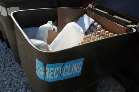 Green recycling bin with cardboard, plastic, and egg cartons placed outdoors on white gravel, promoting eco-friendly waste managementの写真素材
