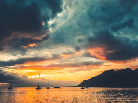Dramatic nature sunset in tropical island lagoon. Yachts silhouette against bright orange blue clouds in sky. Ocean calm water and mountain hill. Wonderful picturesque scene. Majestic travel landscapeの写真素材