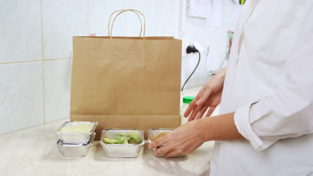 Food delivery service in restaurant. Chef packs lunch boxes to paper bag. Healthy diet daily menu, food meal dishes in a plastic takeaway containers. Woman cook on white kitchen collecting food boxの写真素材