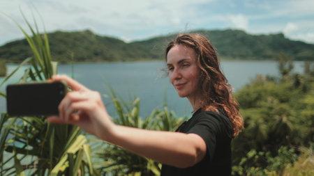 Woman makes selfie mobile photo enjoying tropical jungles and mountain lake landscape. Nature freedom adventure, happy vacation video call, girl tourist outdoors. Travel, tourism, holiday in Polynesiaの写真素材